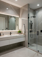 Load image into Gallery viewer, Modern bathroom interior with a large walk-in shower, glass door, floating white vanity, marble-look walls and floor, and a long mirror with built-in lighting.
