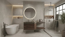 Load image into Gallery viewer, Modern bathroom interior with a freestanding white bathtub, floating wooden vanity with integrated sink and illuminated round mirror, glass shower enclosure, and large window.
